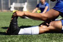 Jogador de futebol vestindo um treinamento de tira de equipe em um campo de esportes ao sol, aquecendo-se esticando as pernas . — Fotografia de Stock