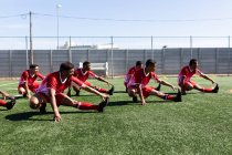 Multi ethnic team of male five a side football players wearing a team strip training at a sports field in the sun, warming up stretching. — Stock Photo