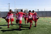 Equipo multiétnico de cinco hombres un lado jugadores de fútbol usando un equipo de entrenamiento de striptease en un campo de deportes en el sol, calentándose de pie en círculo apoyándose mutuamente estiramiento. - foto de stock