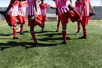 Équipe multiethnique de cinq hommes joueurs de football latéral portant un entraînement de bande d'équipe sur un terrain de sport au soleil, se réchauffant debout en cercle s'étendant. — Photo de stock