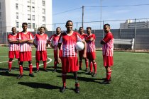 Портрет багатонаціональної команди чоловіків п'ять гравців на боці футболістів у командній стрип-тренінгу на спортивному полі на сонці, що стоїть, тримаючи м'яч посміхаючись камері — стокове фото
