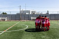 Multi squadra etnica di cinque maschi un giocatore di calcio di lato che indossa un allenamento di striscia di squadra in un campo sportivo al sole, in piedi in huddle motivarsi a vicenda. — Foto stock