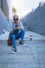 Mixed Race Alternative Frau mit kurzen blonden Haaren unterwegs in der Stadt an einem sonnigen Tag, mit Sonnenbrille und Jeans Latzhose, sitzt auf Stufen mit dem Smartphone. Urbaner digitaler Nomade unterwegs. — Stockfoto