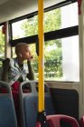 Femme alternative de race mixte avec les cheveux blonds courts dehors et environ dans la ville, assis sur un bus portant des écouteurs sans fil et regardant par la fenêtre. Nomade numérique urbain en déplacement. — Photo de stock