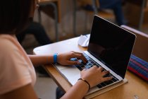 Vue latérale de la section médiane d'une adolescente métisse assise à un bureau en classe à l'aide d'un ordinateur portable — Photo de stock