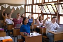 Vista frontal de un grupo multiétnico de alumnos adolescentes sentados en escritorios en un aula y levantando las manos - foto de stock