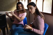 Seitenansicht von zwei kaukasischen Teenager-Mädchen mit langen dunklen Haaren, die vor einem Fenster sitzen, Flöte und Ukulele spielen und in die Kamera schauen — Stockfoto
