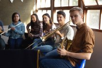 Vue latérale d'un adolescent blanc tenant assis devant une fenêtre dans une salle de classe tenant un cor français avec un groupe multi-ethnique d'adolescents musiciens masculins et féminins assis derrière lui — Photo de stock