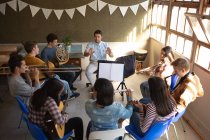 Hochwinkel-Frontansicht eines kaukasischen männlichen Lehrers, der in einem Klassenzimmer sitzt und eine multiethnische Gruppe jugendlicher männlicher und weiblicher Musiker bei einer Schulbandprobe dirigiert — Stockfoto