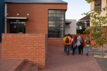 Vista trasera de un grupo multiétnico de estudiantes adolescentes masculinos y femeninos con bolsas de escuela caminando entre edificios en sus terrenos escolares - foto de stock