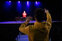 Задній вигляд Кавказького підлітка, який стоїть у залі, де керує підлітком на сцені в порожньому шкільному театрі під час репетицій для виступу. — стокове фото