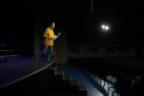 Vista lateral de un adolescente caucásico parado en el escenario sosteniendo un guion en un teatro escolar vacío durante los ensayos para una actuación - foto de stock