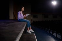 Seitenansicht eines kaukasischen Teenagers, der am Bühnenrand sitzt und in einem leeren Schultheater während der Proben für eine Aufführung ein Drehbuch hält — Stockfoto