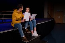 На передньому плані Кавказька дівчинка-підліток і хлопчик сидять на краю сцени з сценаріями і посміхаються один одному в порожній шкільний театр під час репетицій для виступу. — стокове фото
