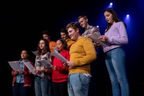 Вид збоку крупним планом багатоетнічна група підлітків-чоловіків і жінок-хореографів, що тримають ноти і співають на сцені шкільного театру під час репетицій для виступу — стокове фото