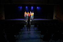 Vista trasera de una mujer caucásica dirigiendo un grupo multiétnico de coristas adolescentes masculinos y femeninos sosteniendo partituras y cantando de pie en el escenario de un teatro escolar durante los ensayos para una actuación - foto de stock