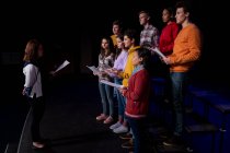 Боковой вид кавказской режиссерки и многоэтнической группы подростков-певчих, стоящих напротив друг друга, держащих ноты и поющих на сцене школьного театра во время репетиций спектакля — стоковое фото