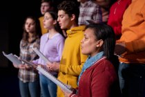 Vista lateral de perto de um grupo multi-étnico de coristas adolescentes e mulheres segurando partituras e cantando em pé no palco de um teatro escolar durante os ensaios para uma performance — Fotografia de Stock