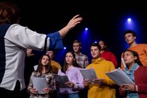 Вид сбоку на кавказскую женщину-дирижера, режиссирующую группу подростков-певцов, держащих ноты и поющих стоя на сцене школьного театра во время репетиций спектакля — стоковое фото