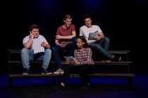 Vista frontal de tres adolescentes caucásicos y una adolescente de raza mixta sosteniendo guiones y sonriendo, sentados en el escenario de un teatro escolar durante los ensayos para una actuación - foto de stock
