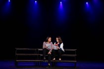 Вид спереди кавказки и смешанной расы девочки-подростки, держащие сценарии, сидящие на сцене школьного театра во время репетиций к спектаклю — стоковое фото