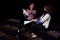 Seitenansicht einer kaukasischen und einer gemischten Rasse Teenager-Mädchen mit Drehbüchern und lachen, die auf der Bühne eines Schultheaters während der Proben für eine Aufführung sitzen — Stockfoto
