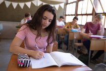 Front view close up of a Caucasian teenage girl sitting at a desk in a school classroom writing in a notebook, with classmates sitting at desks working in the background — Stock Photo