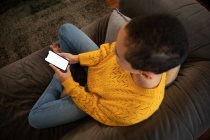 Vista ad alto angolo di una donna mista che si rilassa a casa, seduta su un divano con le gambe incrociate, utilizzando uno smartphone — Foto stock