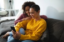 Боковой вид смешанной расы женская пара расслабляется дома, сидя на диване обнимаясь, улыбаясь и глядя на смартфон — стоковое фото