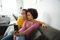 Вид спереду змішаної раси жіноча пара розслабляється вдома, сидячи на дивані разом, один з них тримає смартфон і обидва позують для селфі — стокове фото
