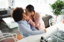 Front view of mixed race female couple relaxing at home, embracing and kissing passionately in their kitchen sitting at the kitchen island — Stock Photo