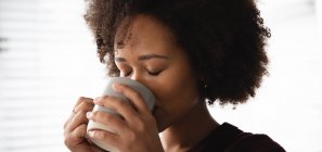 Nahaufnahme einer Frau mit gemischter Rasse, die es sich zu Hause gemütlich macht, eine Tasse Kaffee trinkt, ihn mit beiden Händen hält, mit geschlossenen Augen — Stockfoto