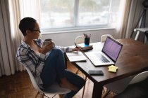 Vue latérale d'une femme métissée assise à une table à la maison, tenant une tasse de café et écrivant dans un carnet, avec un ordinateur portable sur la table devant elle — Photo de stock