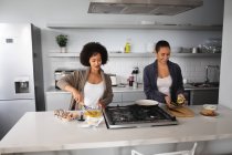 Вид спереду змішаної раси жіноча пара розслабляється вдома, стоячи на кухні, готуючи сніданок разом і посміхаючись разом — стокове фото