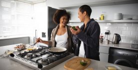 Вид спереду змішаної раси жіноча пара розслабляється вдома, стоїть на кухні, готуючи сніданок разом і сміючись. Вони б'ють яйця і тримають авокадо — стокове фото