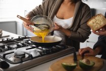 Frontansicht einer Frau, die in ihrer Küche frühstückt, geschlagene Eier aus einer Schüssel in eine Pfanne auf dem Herd gießt, ihr Partner neben ihr hält ein Stück Butterbrot mit einer aufgeschnittenen Avocado auf einem Schneidebrett vor sich. — Stockfoto