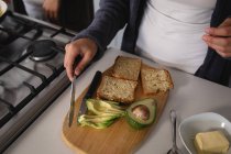 Visão frontal de alto ângulo no meio da seção da mulher relaxando em casa, preparando café da manhã na cozinha, corte de abacate e pão de manteiga em uma tábua de cortar — Fotografia de Stock