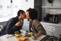 Vorderansicht eines gemischten Rassenpaares, das es sich zu Hause gemütlich macht, auf Stühlen auf der Kücheninsel sitzt, frühstückt und küsst und Tassen Kaffee hält — Stockfoto