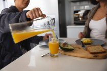 Seitenansicht Mittelteil des gemischten Rennens weibliches Paar entspannt zu Hause, sitzt an einem Tisch in der Küche beim Frühstück, einer gießt ein Glas Orangensaft aus einem Krug — Stockfoto