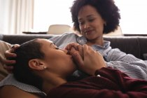 Side view close up of a mixed race female couple relaxing at home in the living room on the couch together in the morning, one woman lying with her head on the lap of her seated partner, holding and kissing her hand — Stock Photo