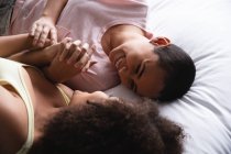 Side view close up view of a mixed race female couple relaxing at home in the bedroom together in the morning, lying on the bed in their nightclothes, holding hands and looking at each other smiling — Stock Photo