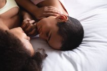 Großaufnahme eines gemischten weiblichen Paares, das es sich morgens zu Hause im Schlafzimmer gemütlich macht, einander gegenüber auf dem Bett liegend und Händchen haltend — Stockfoto
