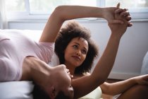 Front view close up of a mixed race female couple relaxing at home in the bedroom in the morning, one lying on her back on the bed and the other sitting beside her on the floor, holding hands and smiling — Stock Photo