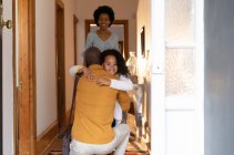 Вид спереди на молодую афроамериканскую девушку, обнимающую своего отца, когда он стоит на коленях в коридоре дома и улыбается через плечо, а ее мать стоит позади нее, улыбаясь — стоковое фото