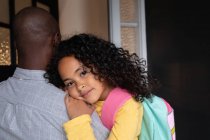 Nahaufnahme eines jungen afroamerikanischen Mädchens mit langen lockigen Haaren, das einen Rucksack trägt, in die Kamera lächelt und sich auf die Schulter ihres Vaters stützt, als er sie auf dem Flur zu Hause hält — Stockfoto