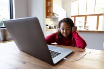 Vue de face d'une jeune afro-américaine à la maison, assise à la table du dîner à écouter les yeux fermés et écouteurs allumés, branchée sur un ordinateur portable sur la table devant elle et souriant — Photo de stock