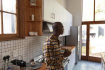 Vue latérale d'un Afro-Américain à la maison, debout dans la cuisine portant une chemise à carreaux, tenant une tasse de café et regardant par la fenêtre vers le jardin — Photo de stock
