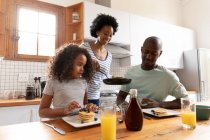 Front view of a young African American girl and her father at home in the kitchen in the morning, sitting at the kitchen island, the mother standing behind serving them pancakes from a frying pan — Stock Photo
