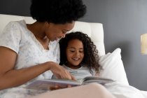 Front view close up of an African American woman and her young daughter relaxing in the bedroom, sitting up in bed and reading a book together — Stock Photo
