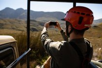 Vue latérale de l'homme caucasien profitant du temps dans la nature, dans un équipement de tyrolienne assis dans une voiture, prendre des photos avec smartphone par une journée ensoleillée dans les montagnes — Photo de stock